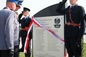 Komendant Wojewódzki stoi przy obelisku i czyta nazwiska pomordowanych policjantów. Przy obelisku policjant i policjantka oddają honor w mundurze z epiki 20-lecia międzywojennego.