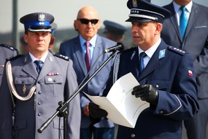 Dyrektor Biura Historii i Tradycji przy mikrofonie trzyma teczkę. W tle oficer Policji.