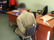 Zatrzymany mężczyzna siedzi na krześle obok biurka z kajdankami założonymi na ręce trzymane z tyłu. Ma zamazaną twarz.
