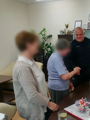 Zastępca Komendanta Miejskiego Policji w Poznaniu komisarz Piotr Gruszka ściska dłoń seniorki, która odzyskała swoje pieniądze. Na pierwszym planie córka starszej kobiety. Obie panie mają zamazane twarze.