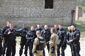 Policjanci w czarnym umundurowaniu ćwiczebnym stoją i patrzą się na dwóch instruktorów prowadzących szkolenie
