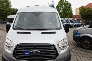 Przód srebrnego forda transita, przez szybę widać siedzących w nim dwóch policjantów, w tle radiowozy,.