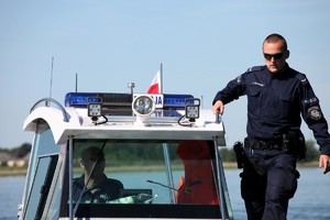 Policyjna motorówka płynie po jeziorze,  a na jej boku stoi policjant w mundurze i okularach słonecznych