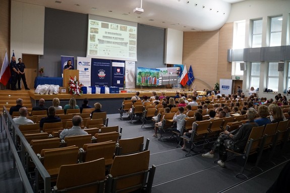 Sala urzędu, w której odbyła się gala, w rzędach siedzą dzieci, nauczyciele, na scenie policjantka prowadząca galę i w tle banery z logami instytucji - organizatorów, patronów konkursu