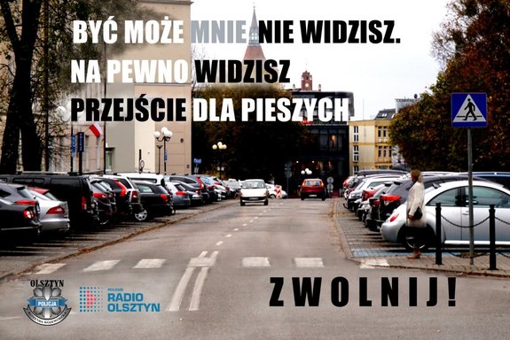 Plakat kampanii, na którym widoczna jest ulica w mieście, jadący samochód i piesza stojąca przy przejściu