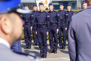 Uczestnicy uroczystego ślubowania nowo przyjętych policjantów. Policjanci stojący w szeregu na tle radiowozów.