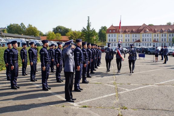 Uczestnicy uroczystego ślubowania nowo przyjętych policjantów. Przemarsz Pocztu Sztandarowego przed policjantami stojącymi w szeregu.