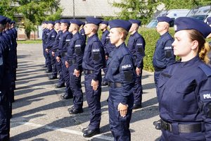 Uczestnicy uroczystego ślubowania nowo przyjętych policjantów. Na pierwszym planie policjanci stojący w szeregu, w tle za żywopłotem dwa stojące radiowozy.