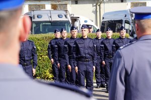 Uczestnicy uroczystego ślubowania nowo przyjętych policjantów. Na pierwszym planie policjanci stojący w szeregu na tle żywopłotu i  dwóch radiowozów. Na drugim planie dwóch oficerów policji stojących tyłem.