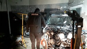 Fotografia kolorowa. Przedstawia wnętrze garażu, a w nim trzy odzyskane samochody.  Jeden z pojazdów jest częściowo rozmontowany, obok niego stoi policjant..