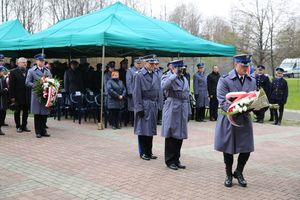 Fotografia kolorowa. Na zdjęciu policjanci oraz zebrani goście składający kwiaty przed grobem.