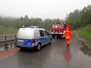 Zdjęcie przedstawia z lewej: policyjny radiowóz, następnie idącego tyłem strażaka na tle wozu strażackiego