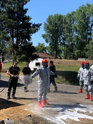 Strażacy w ramach ćwiczeń polewają wodą osoby w kombinezonach ochronnych