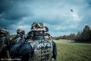 Zdjęcie kolorowe. Uczestnicy szkolenia na lądowisku oczekują, na dalszym planie śmigłowiec w powietrzu