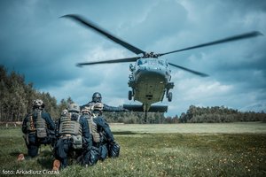 Zdjęcie kolorowe. Uczestnicy szkolenia na lądowisku oczekują, na dalszym planie śmigłowiec w powietrzu