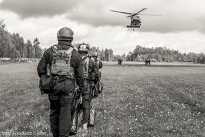 Zdjęcie czarno-białe. Uczestnicy szkolenia na lądowisku oczekują, na dalszym planie śmigłowiec w powietrzu