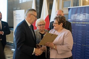 Zdjęcie kolorowe. Zdjęcie przedstawia wręczenie wyróżnienia jednej z uczestniczek konferencji za działalność społeczną, przez Stanisława Szweda - Sekretarza Stanu w Ministerstwie Rodziny, Pracy i Polityki Społecznej, oraz poseł na Sejm RP Marie Nowak.