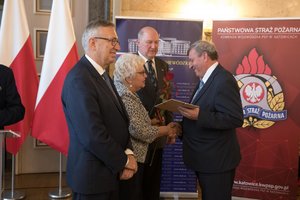 Zdjęcie kolorowe. Zdjęcie przedstawia wręczenie wyróżnienia za działalność społeczną uczestnikowi konferencji. Wyróżnienie wręcza poseł na Sejm RP Maria Nowak, Stanisław Szwed - Sekretarz Stanu w Ministerstwie Rodziny, Pracy i Polityki Społecznej, oraz wicewojewoda śląski Jan Chrząszcz.