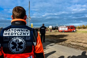 Na pierwszym planie ratownik medyczny. W tle policjant i służby ratunkowe przy przewróconym autobusie.