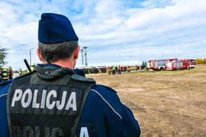 Na pierwszym planie policjant stojący plecami. W tle działania służb ratunkowych przy przewróconym autobusie.