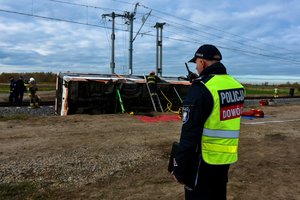 Na pierwszym planie policjant z radiotelefonem. W tle strażacy przy przewróconym autobusie.