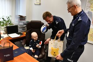 Kubuś siedzący za biurkiem na miejscu naczelnika, obok niego Zastępca Komendanta Wojewódzkiego Policji w Katowicach, insp. Mariusz Krzystyniak oraz naczelnik. Wszyscy oglądają wspólnie prezenty, które otrzymało dziecko