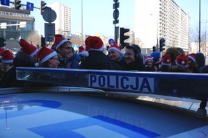 na pierwszym planie oznakowany policyjny radiowóz, w tle młodzież w czapkach Świętego Mikołaja