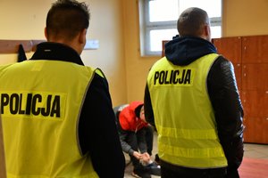 dwóch policjantów ubranych w kamizelki odblaskowe z napisem POLICJA. Pomiędzy nimi widać zatrzymanego mężczyznę