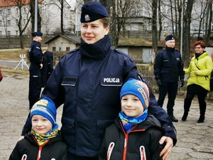 Policjantka trzyma za ramiona dwóch chłopców stojących po obydwu jej stronach. W tle inni policjanci ze swoimi bliskimi.