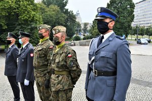 Przedstawiciele służb mundurowych stoją w szeregu przed pomnikiem