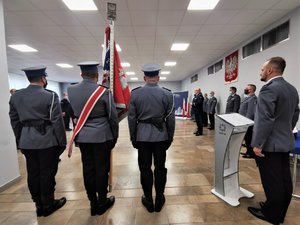 Poczet Sztandarowy stojący tyłem, obok policyjny spiker. W tle przemawia Komendant Wojewódzki Policji w Katowicach.