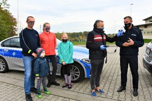 Umundurowany policjant udziela informacji dla radia, obok dziennikarza trzymającego mikrofon stoi kobieta, mężczyzna i dwójka dzieci. Wszyscy mają odblaskowe bransoletki. Z tyłu zaparkowany policyjny radiowóz.