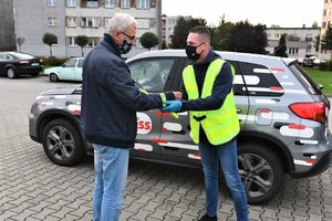 Dziennikarz wręcza mężczyźnie odblaskową bransoletkę. W tle samochód z logo Radia Express.