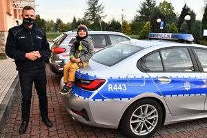 Na zdjęciu policjant z dzieckiem siedzącym na klapie bagażnika oznakowanego radiowozu. Dziecko się uśmiecha.