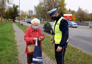 Umundurowany policjant rozmawia na chodniku z kobietą, której wręczył odblaskową torbę. W tle policyjny radiowóz