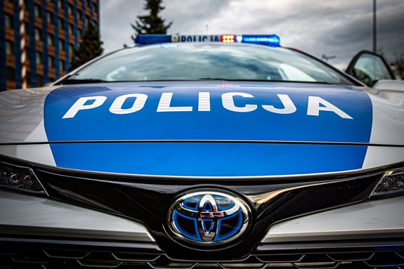 Zdjęcie przodu oznakowanego radiowozu, widoczny napis Policja na masce samochodu. Sygnały świetlne są włączone. W tle niebo i fragment budynku komendy wojewódzkiej.