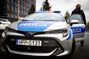 Zdjęcie przodu oznakowanego radiowozu, drzwi od strony kierowcy są otarte, stoi przy nich umundurowany policjant. Sygnały świetlne są włączone.