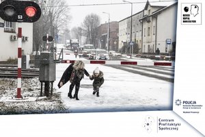 Fotografia przedstawia dorosłą kobietę przechodzącą z dzieckiem pod zamkniętym szlabanem przejazdu kolejowego.