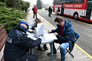 Umundurowany policjant oraz dwie inne osoby siedzą przy stole i wypełniają ankiety przed oddaniem krwi. W tle stoi autobus, w którym pobierana jest krew.