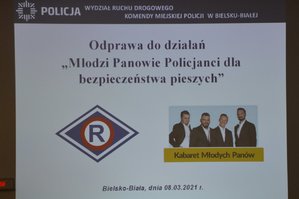 Slajd z prezentacji multimedialnej, a na niej, w górnej części logo Policji oraz napis Wydział Ruchu Drogowego Komendy Miejskiej Policji w Bielsku-Białej, pod spodem napis Odprawa do działań &quot;Młodzi Panowie Policjanci dla bezpieczeństwa pieszych&quot; oraz zdjęcie członków kabaretu, logo ruchu drogowego. Na dole widnieje napis z miejscowością oraz datą.