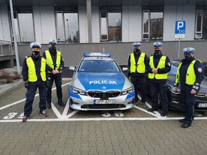 Grupowe zdjęcie kabareciarzy oraz policjantów. Wszyscy są ubrani w policyjne mundury i stoją przy oznakowanym radiowozie.