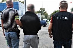 Dwaj nieumundurowani policjanci z wydziału kryminalnego prowadzą zatrzymanego mężczyznę w kierunku radiowozu.