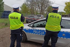 Policjanci w kamizelkach odblaskowych stoją tyłem, a w tle stoi radiowóz policyjny.