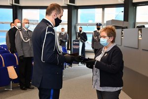 Zdjęcie kolorowe. Widoczni uczestnicy uroczystości na auli, Komendant Wojewódzki Policji w Katowicach wręcza wyróżnienie na ręce P. Teresy Brackiej