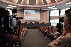 Zdjęcie kolorowe. Briefing prasowy na auli, widoczni uczestnicy wydarzenia oraz policjant, który zabiera głos na mównicy