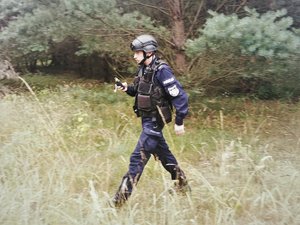 Zdjęcie kolorowe. Widoczni umundurowani policjanci prowadzący poszukiwania w terenie leśnym