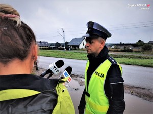 Zdjęcie kolorowe. Widoczny umundurowany policjant udzielający wyiadu dziennikarzom