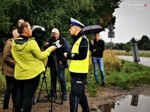 Zdjęcie kolorowe. Widoczny umundurowany policjant udzielający wyiadu dziennikarzom