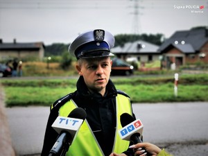 Zdjęcie kolorowe. Widoczny umundurowany policjant udzielający wyiadu dziennikarzom