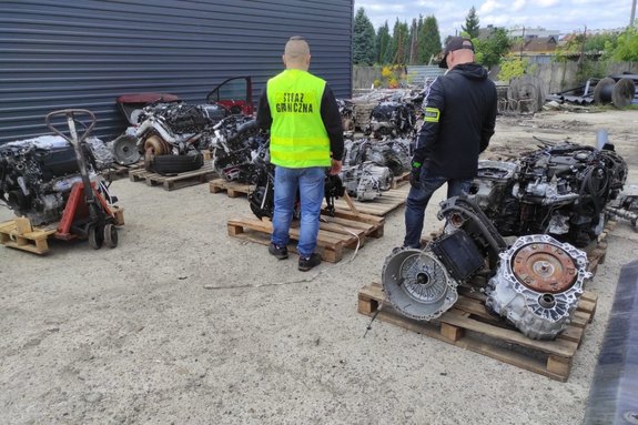 Zdjęcie kolorowe. Widoczne częscie samochodowe składowane na placu oraz policjant i funkcjonariusz strazy granicznej